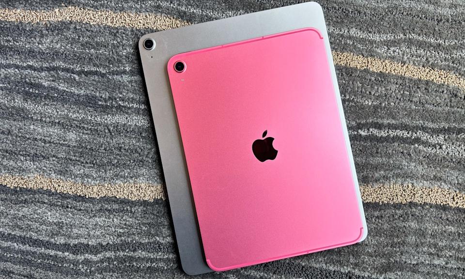 A pink 11-inch Apple iPad rests on top of a silver 13-inch iPad Air. Both tablets are face down on a gray carpet with beige and white streaks.