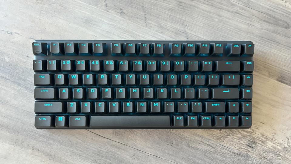 A black wireless keyboard with light blue RGB backlighting, the Alienware Pro Wireless Gaming Keyboard, sits on top of a brown wooden table.