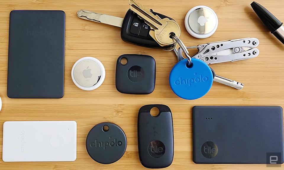 An assortment of bluetooth trackers arranged in a grid on a wooden background. Trackers include black Tile trackers in various shapes, two silver and white AirTag trackers and a round blue Chipolo tracker attached to a set of keys with a multitool key chain.